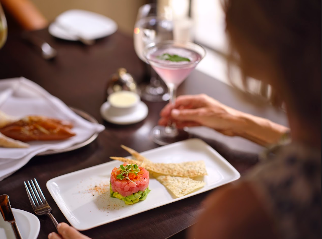 Plated gourmet meal with cocktail on a restaurant table, viewed beside a diner’s hands.
