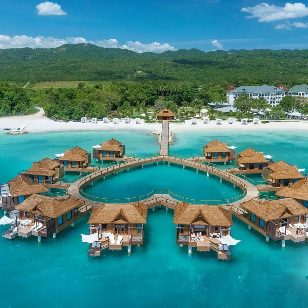 Aerial view of overwater bungalows surrounding a circular pool in bright turquoise lagoon with beach and mountains nearby