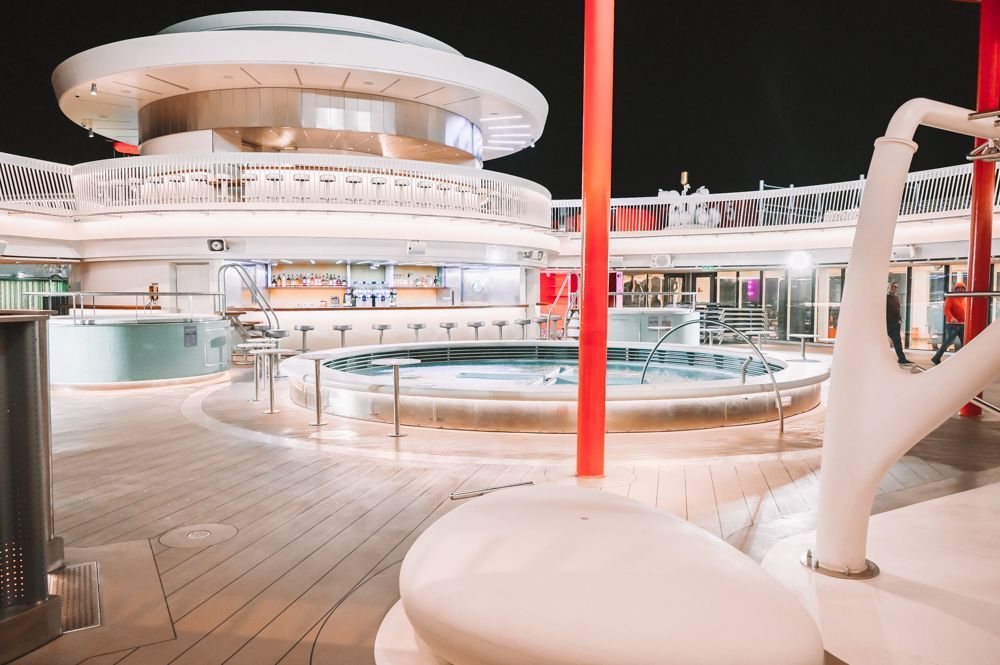 Modern cruise ship pool deck at night with lit circular pool and white lounge area