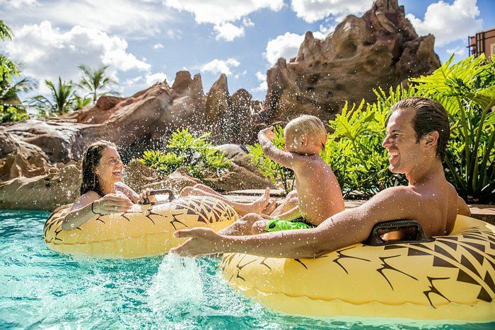 Three people floating in yellow tubes in a turquoise pool with rocky cliffs and palm trees
