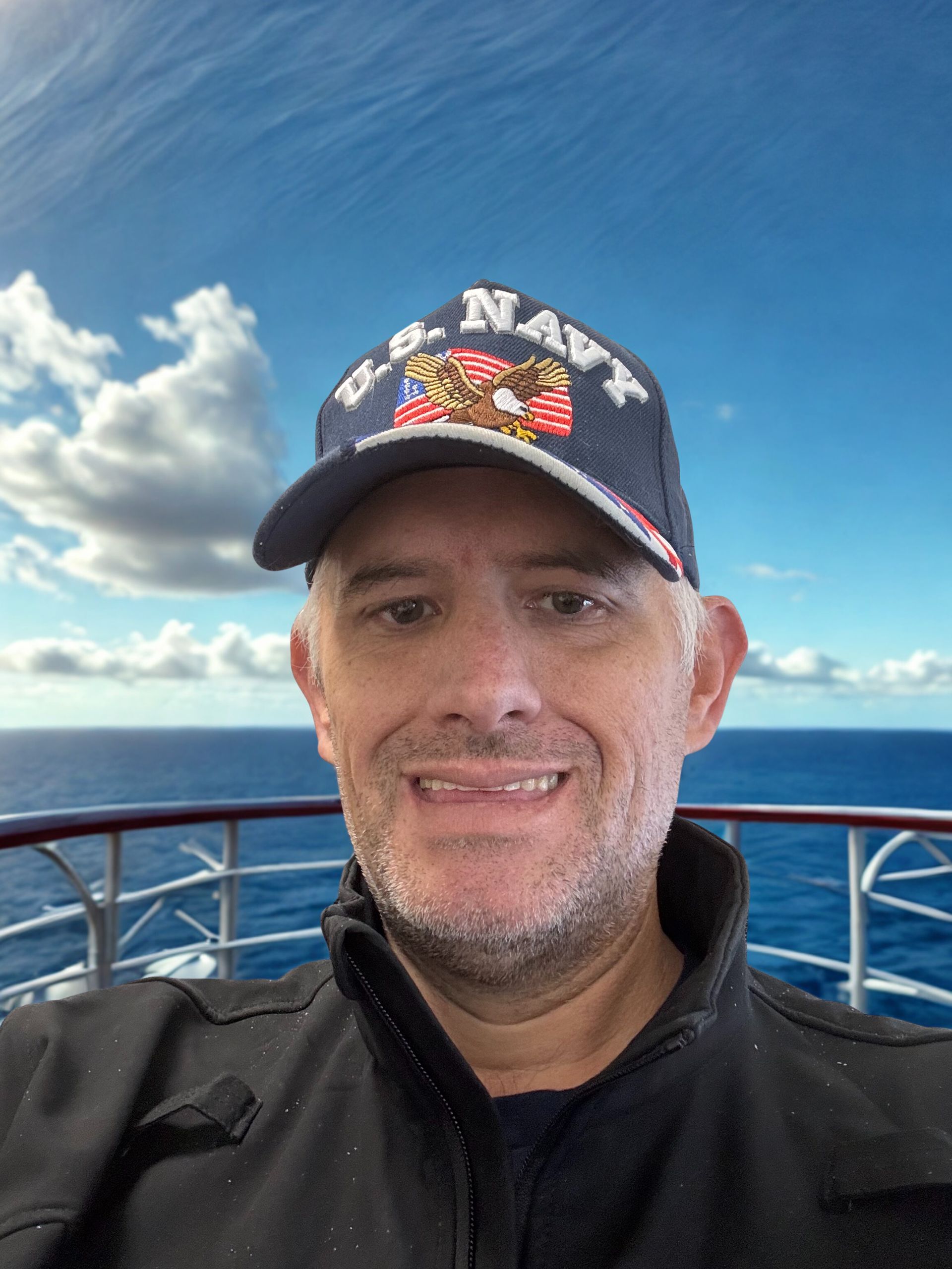 Man in a navy cap smiling on a boat deck with blue ocean and cloudy sky behind him