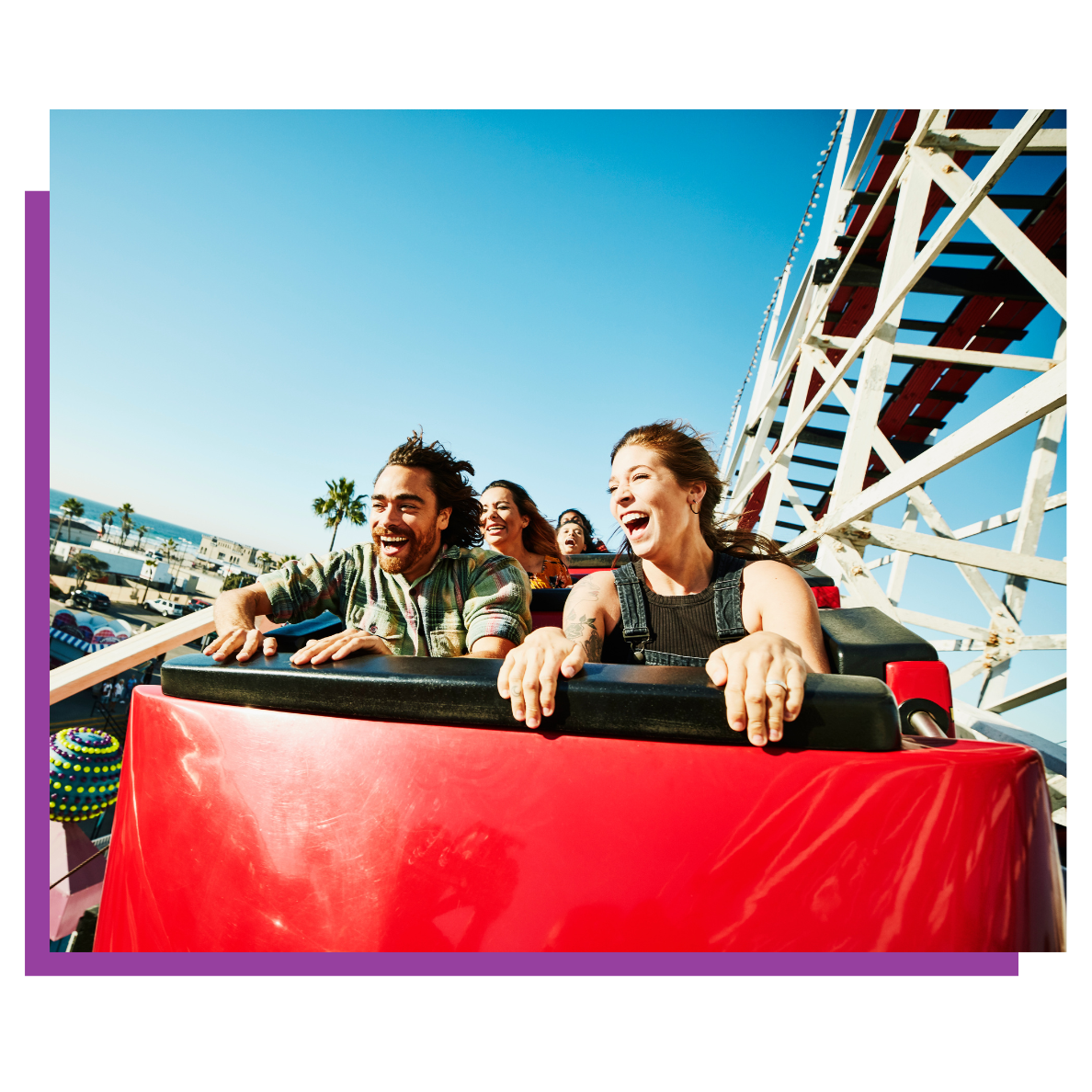 People riding a red roller coaster car, smiling at an amusement park under a bright blue sky.