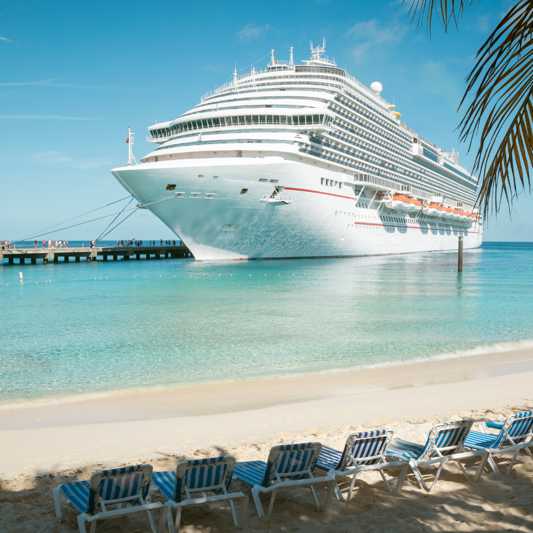 Large white cruise ship anchored in turquoise water beside a sandy beach with lounge chairs under palm fronds