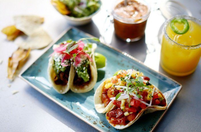 Tacos on a blue plate with salsa, lime, and chips, beside a glass of orange juice