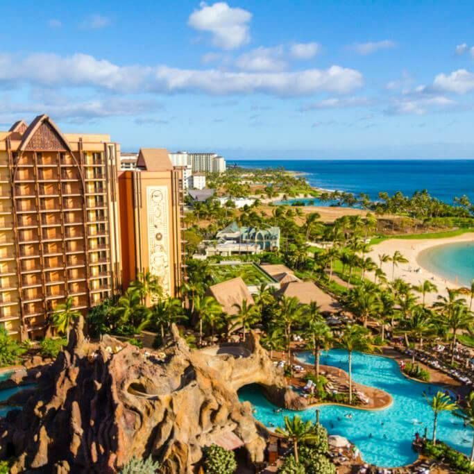 Tropical resort towers beside palm pools, rocky lagoon, and a turquoise ocean under a blue sky.