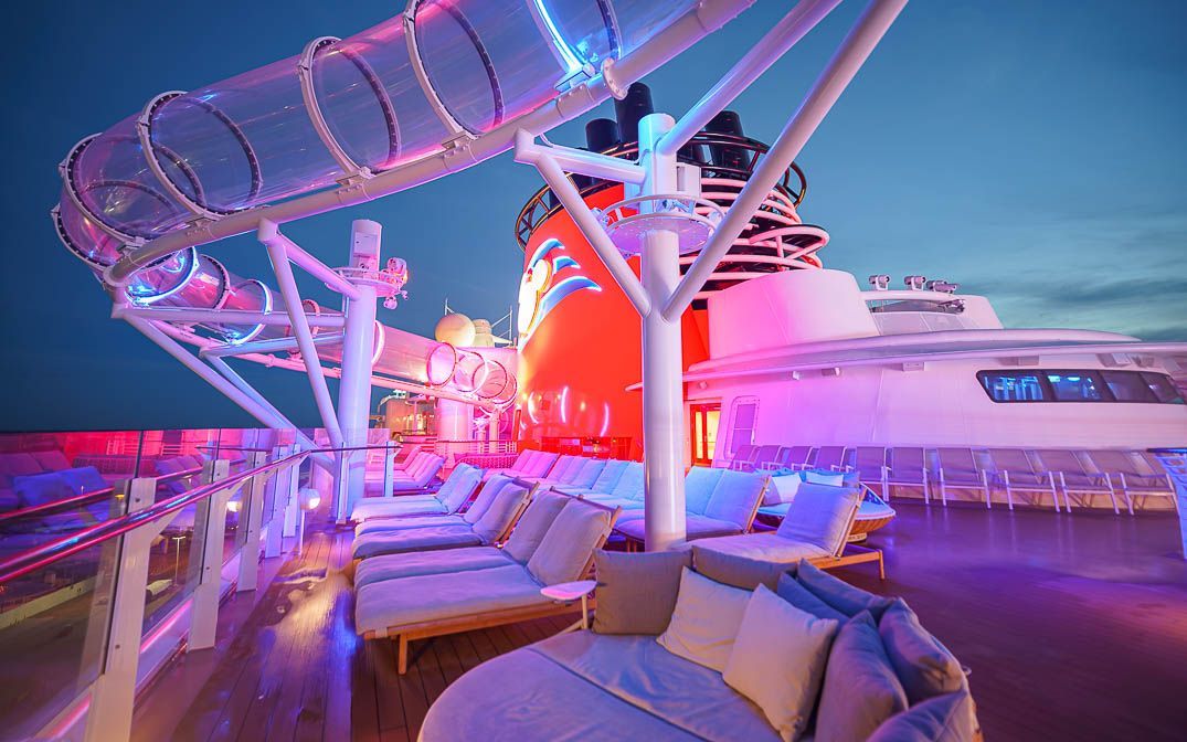 Luxury cruise ship deck at night with lounge chairs, pink lighting, and a water slide overhead