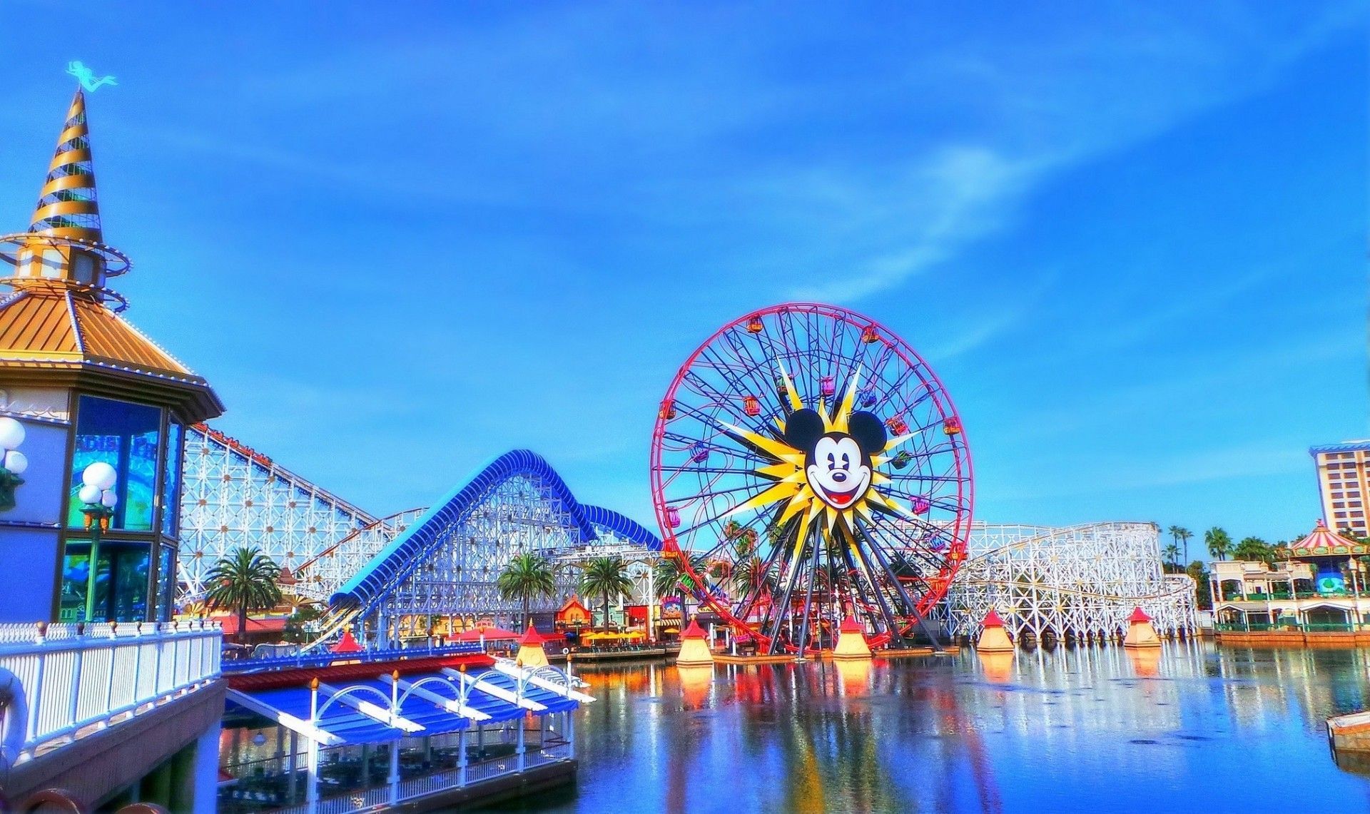 Colorful amusement park with a Mickey-shaped Ferris wheel reflected on calm water under a bright blue sky