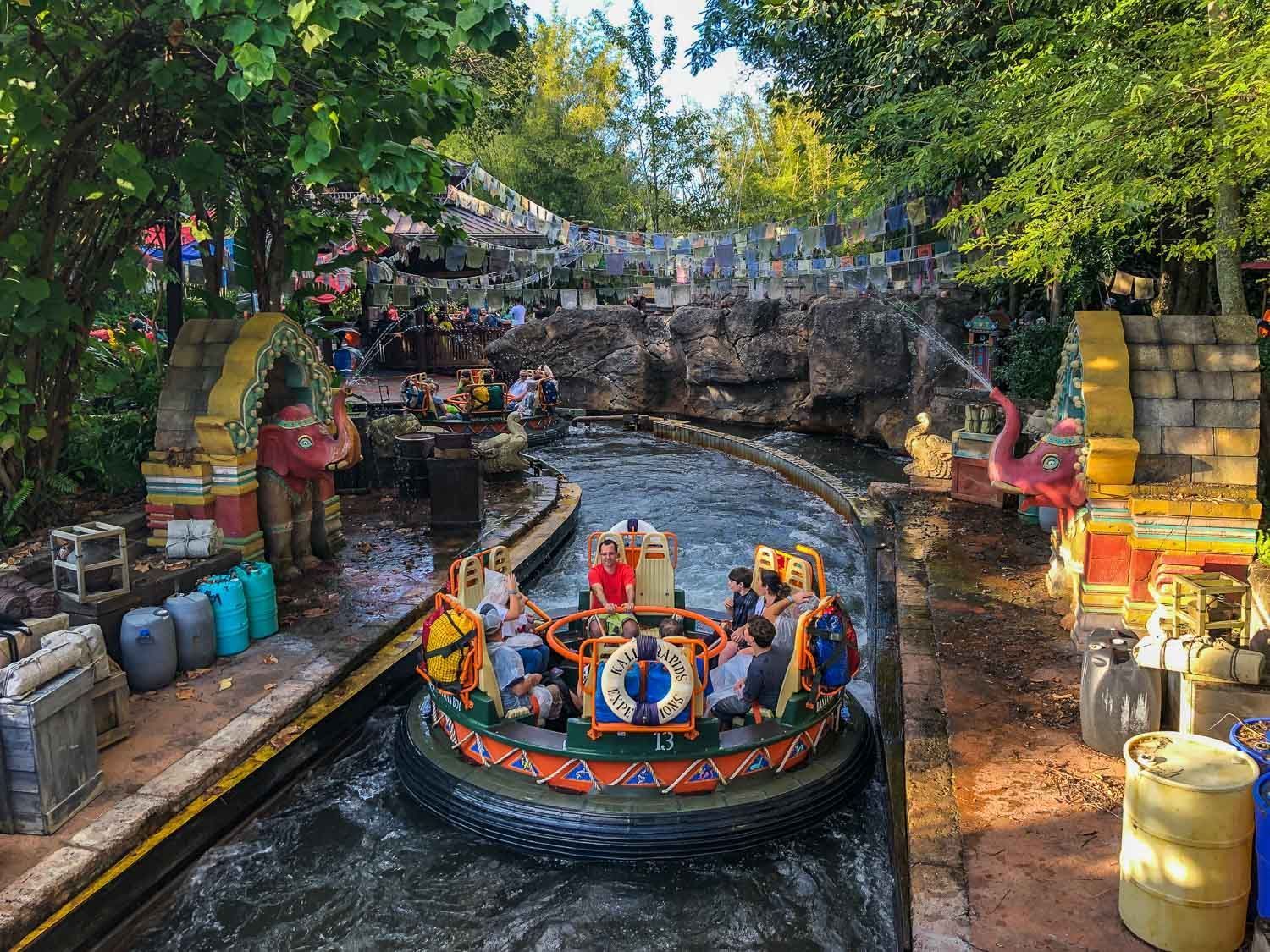 Six people ride a colorful raft on a river in a lush, rocky theme park tunnel.