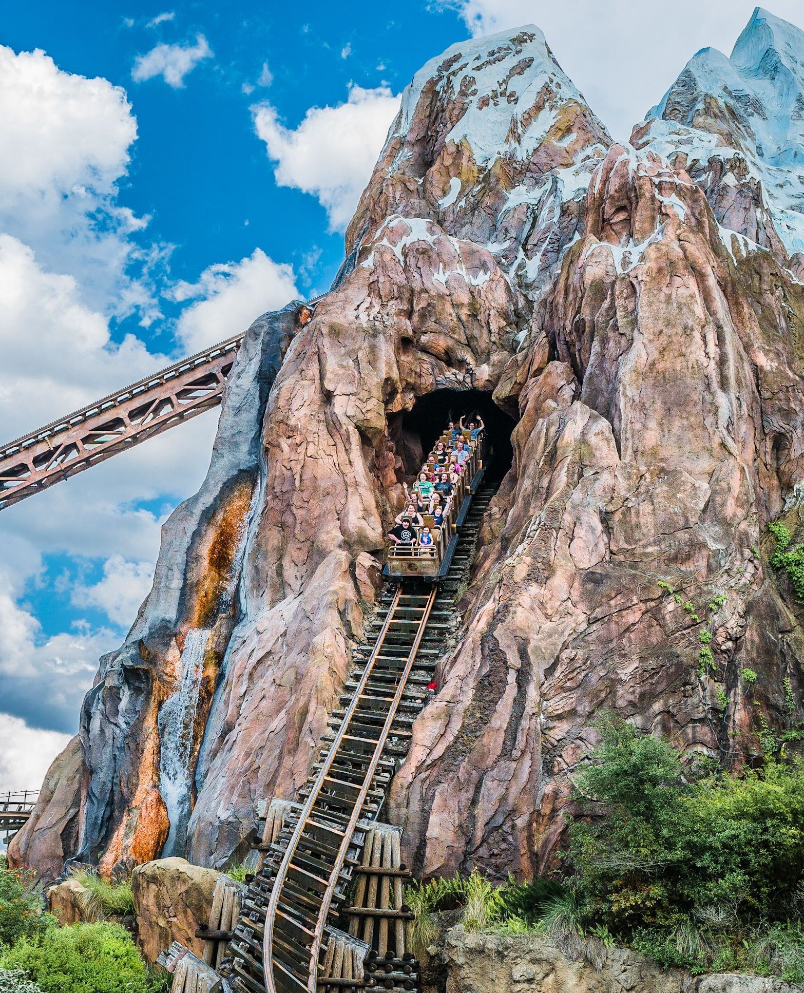 Steep mountain themed ride with a train emerging from a rocky tunnel against a blue sky