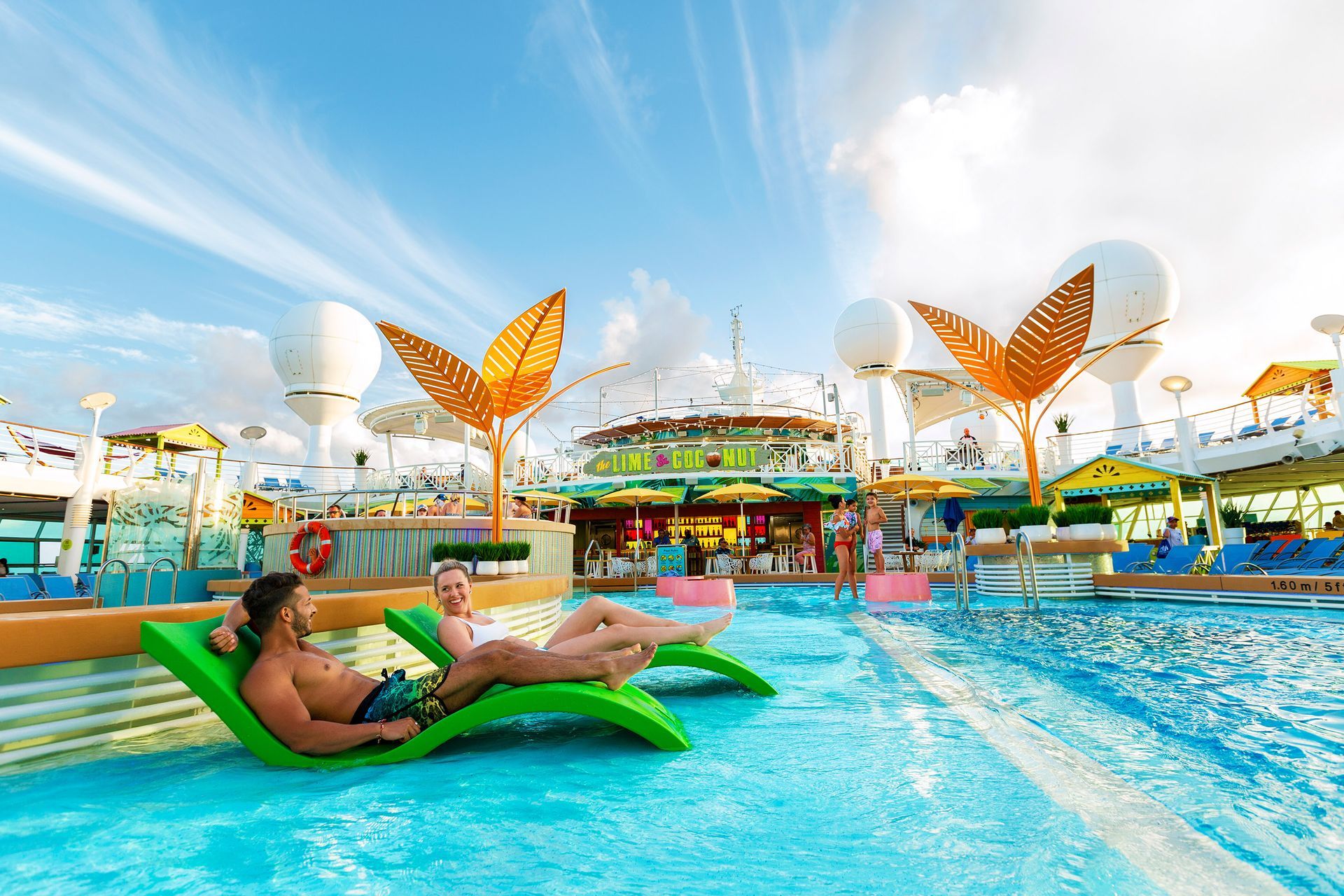 People lounging on green floats in a bright blue resort pool with butterfly sculptures and a sunny sky