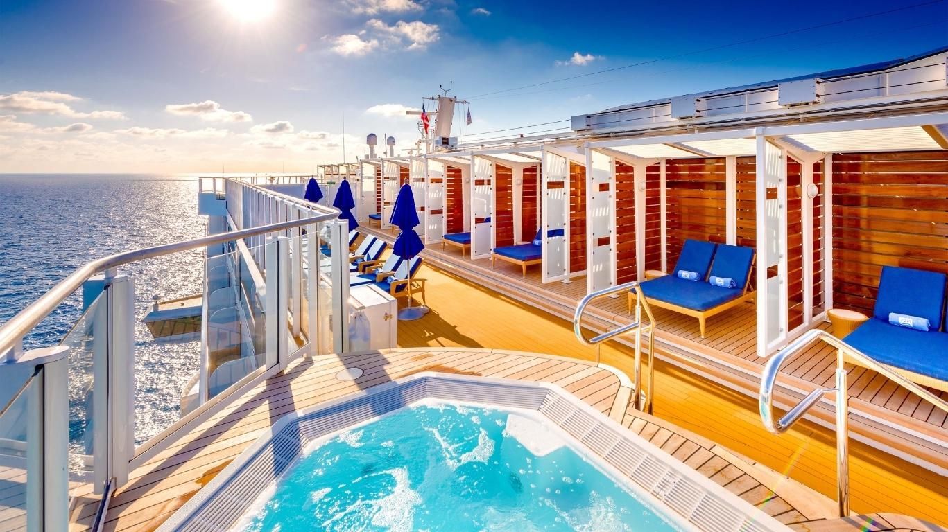 Cruise ship deck with blue lounge chairs, hot tub, and ocean view at sunset