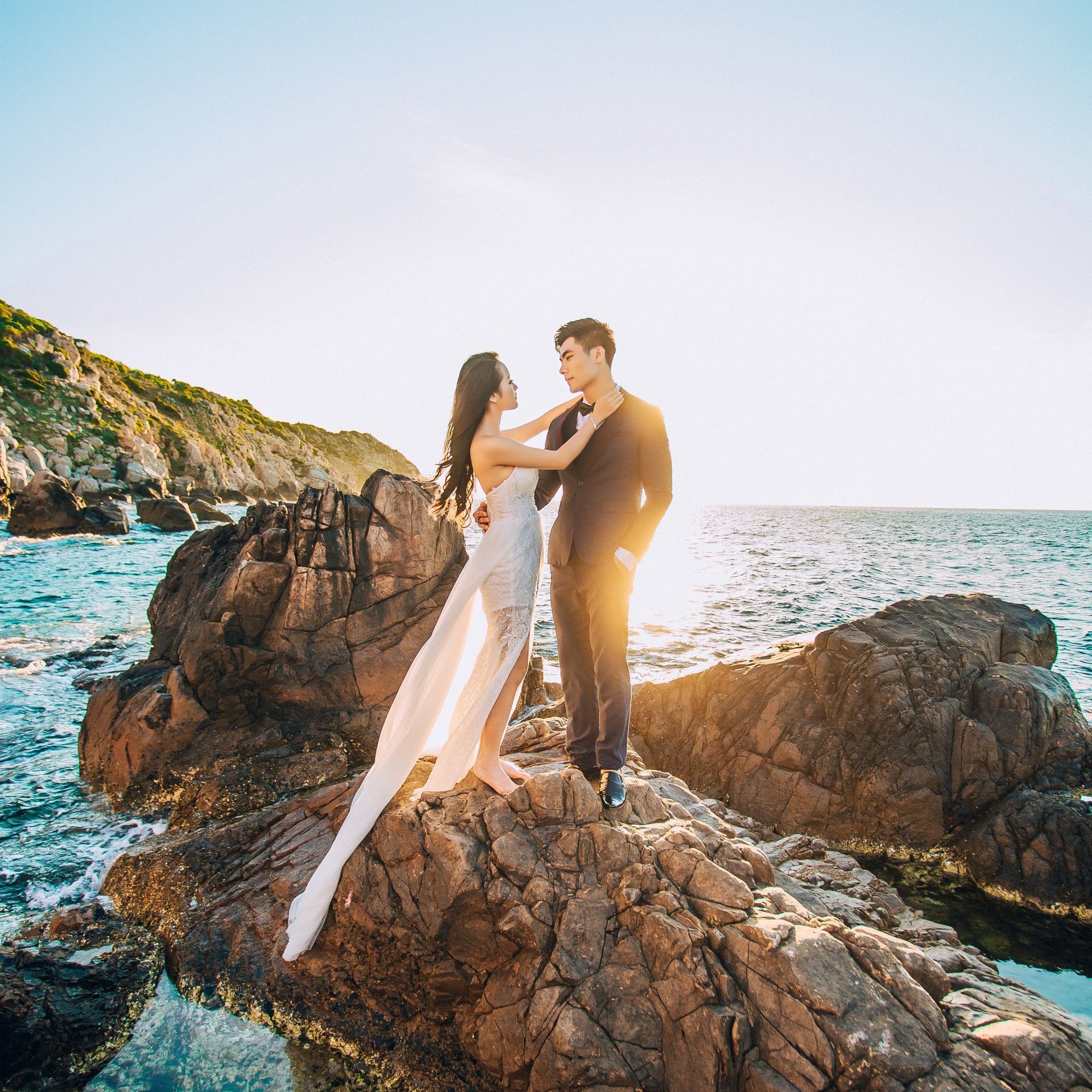 Couple standing on rocky shore at sunset, with ocean waves and bright sky behind them