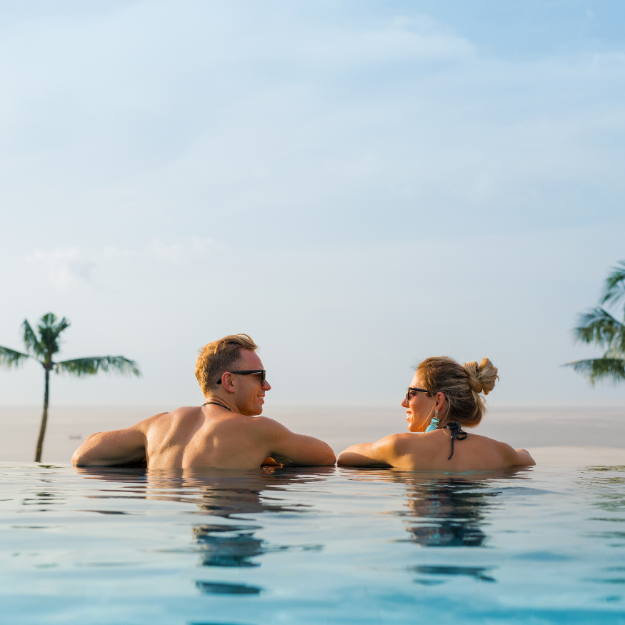 Two people relaxing in an infinity pool, looking out over a calm ocean at sunrise.