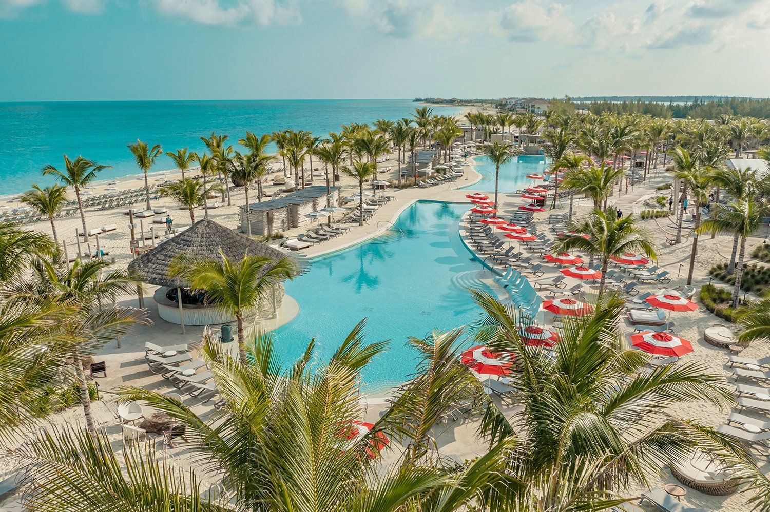 Tropical resort pool beside a sandy beach, lined with red lounge chairs and palm trees under a blue sky