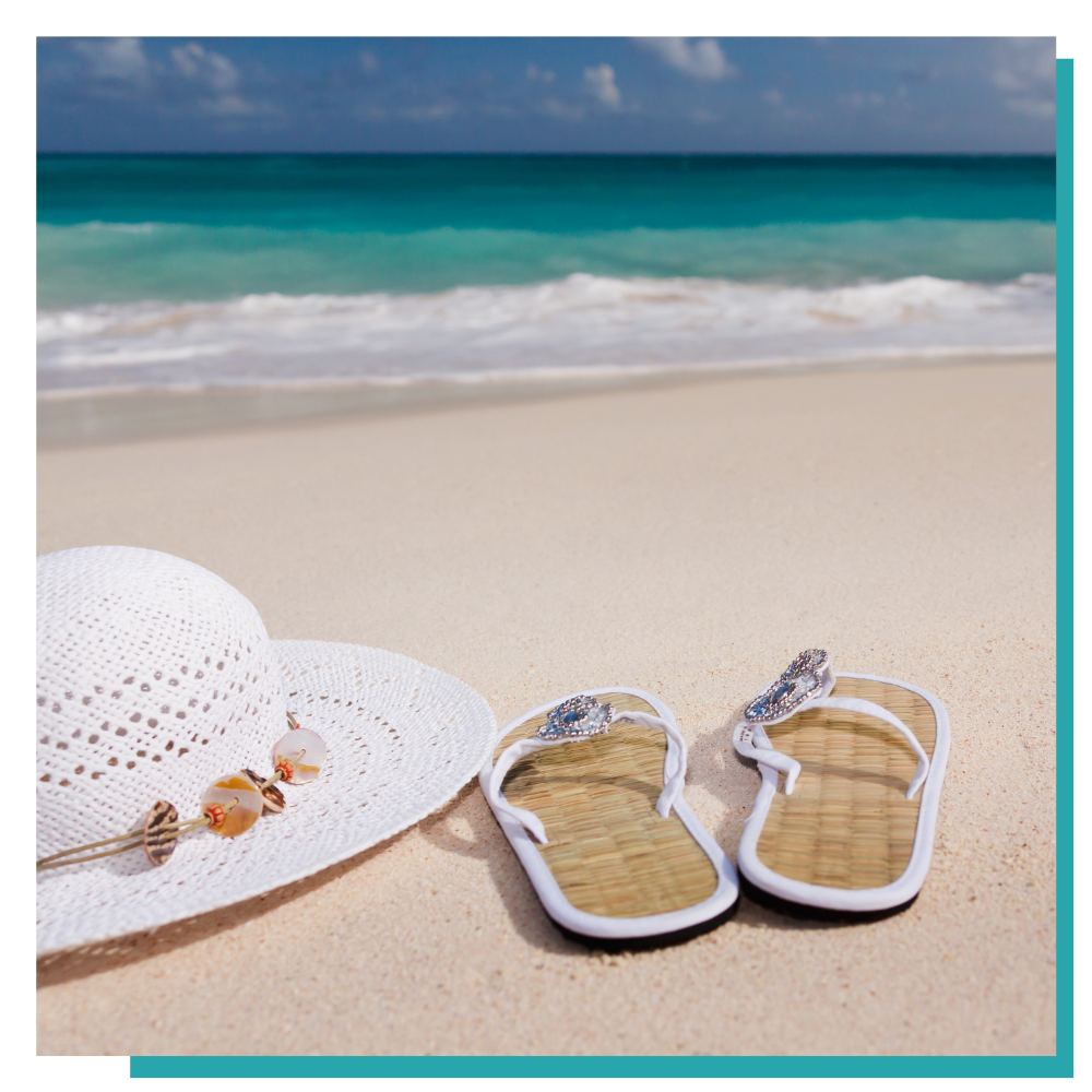White sunhat and sandals on a sandy beach with turquoise ocean waves in the background