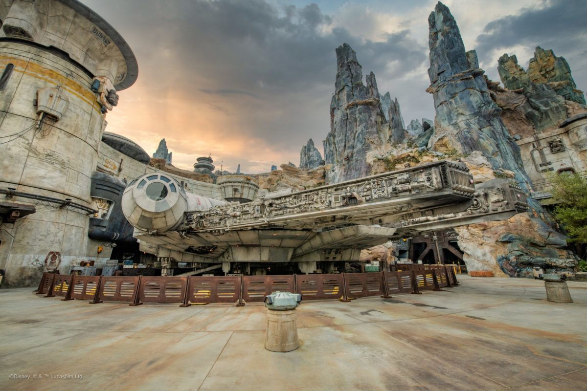 Weathered spaceship in a rocky sci-fi outpost at sunset, with tall jagged spires and a round building.