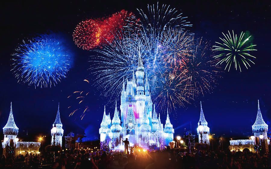 Fireworks burst over a glowing castle at night, with blue, red, and green fireworks above the lit skyline.