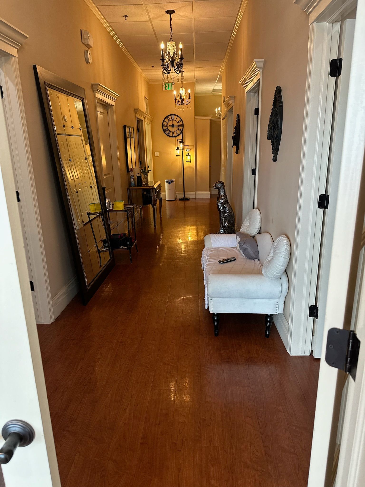 A hallway with warm wooden floors, a large leaning mirror on the left, and a white cushioned bench on the right.