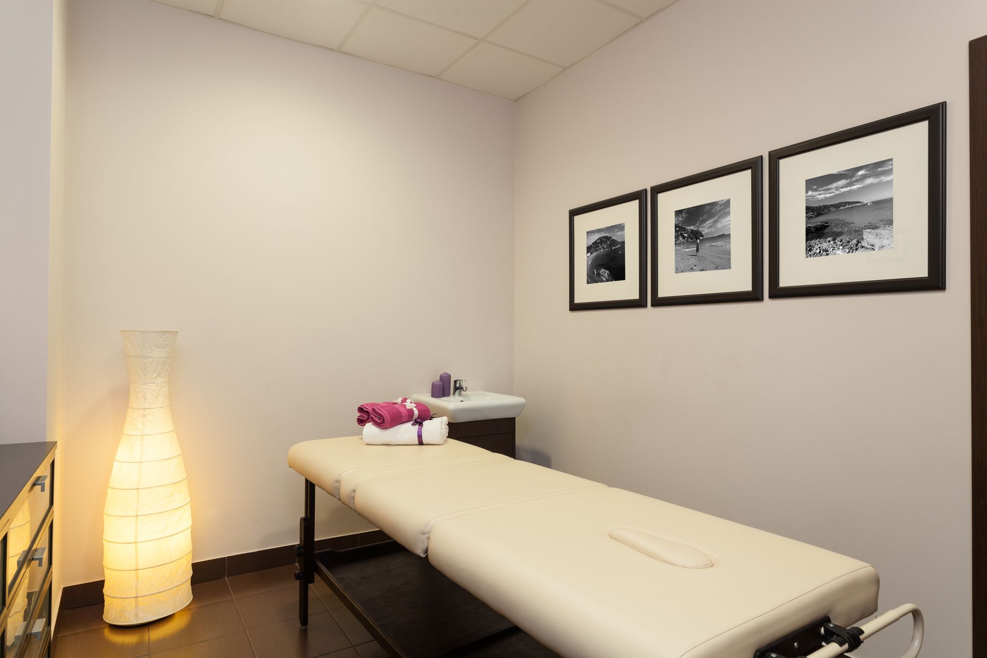 A treatment room featuring a massage table with folded towels, a tall floor lamp, and three framed pictures on the wall.