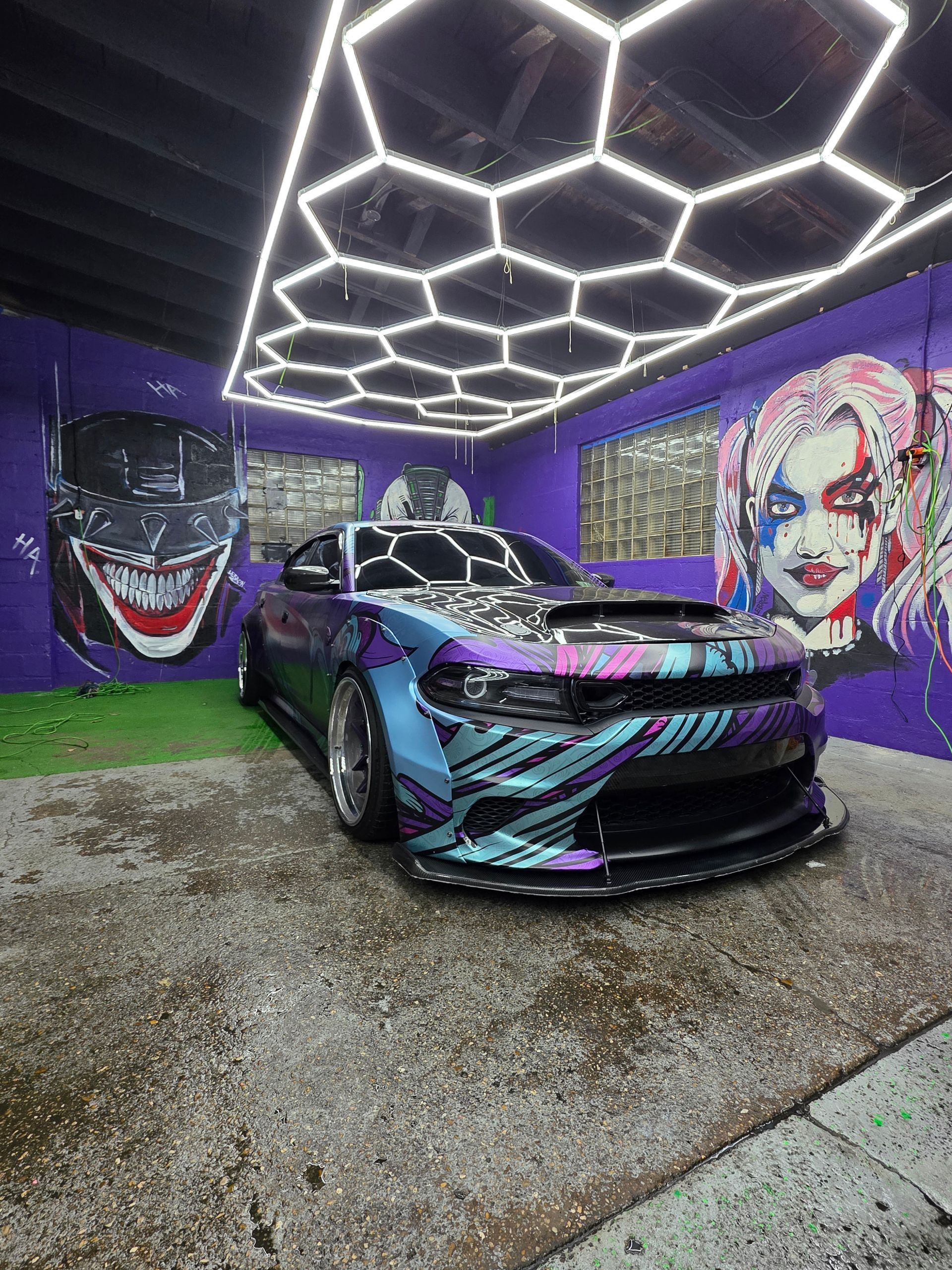 Sports car with purple, blue, and pink wrap parked in a neon-lit garage with Joker murals