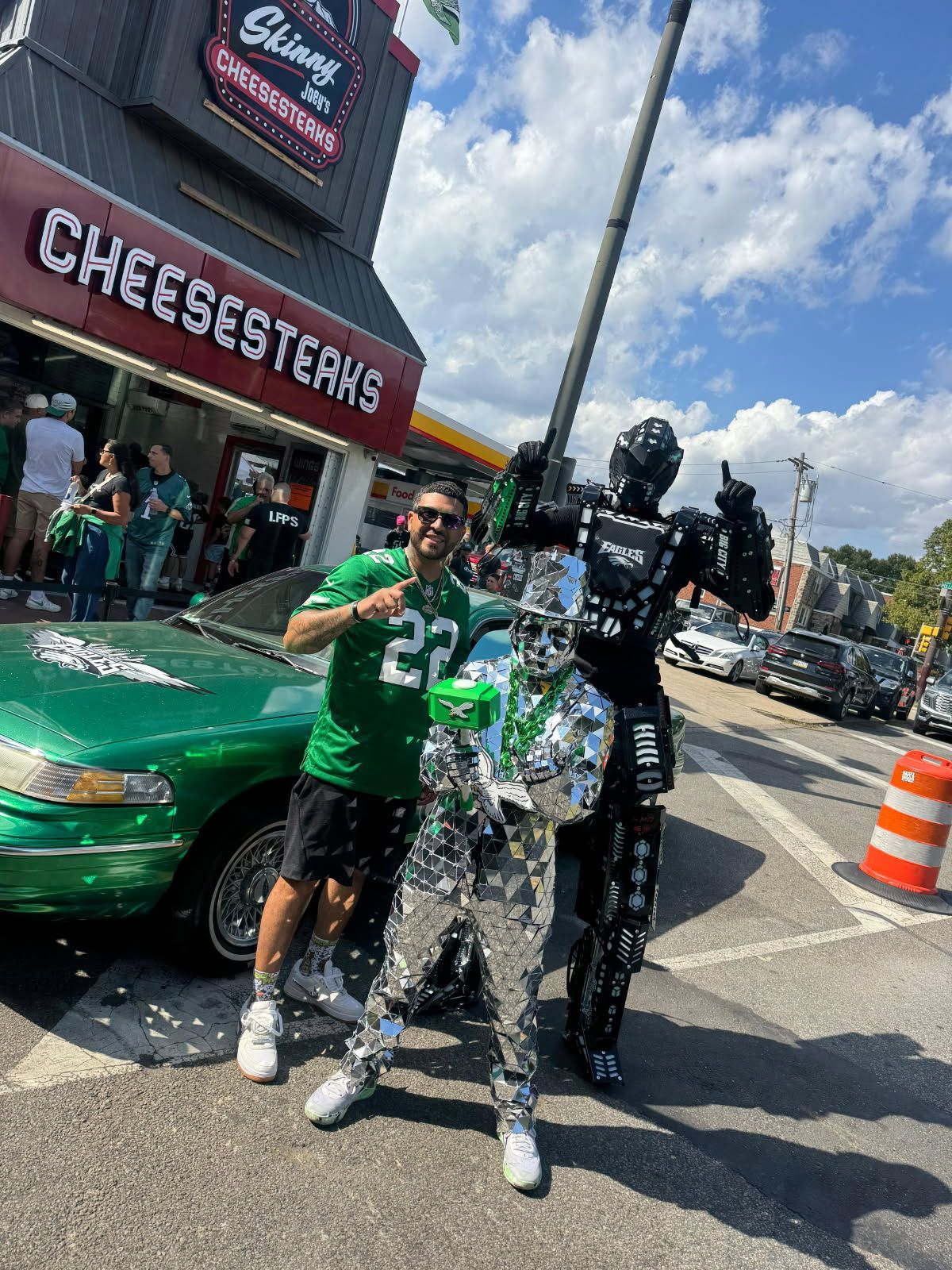 Two people posing beside a green car outside Cheesesteak’s, one in a black robot costume holding a trophy.