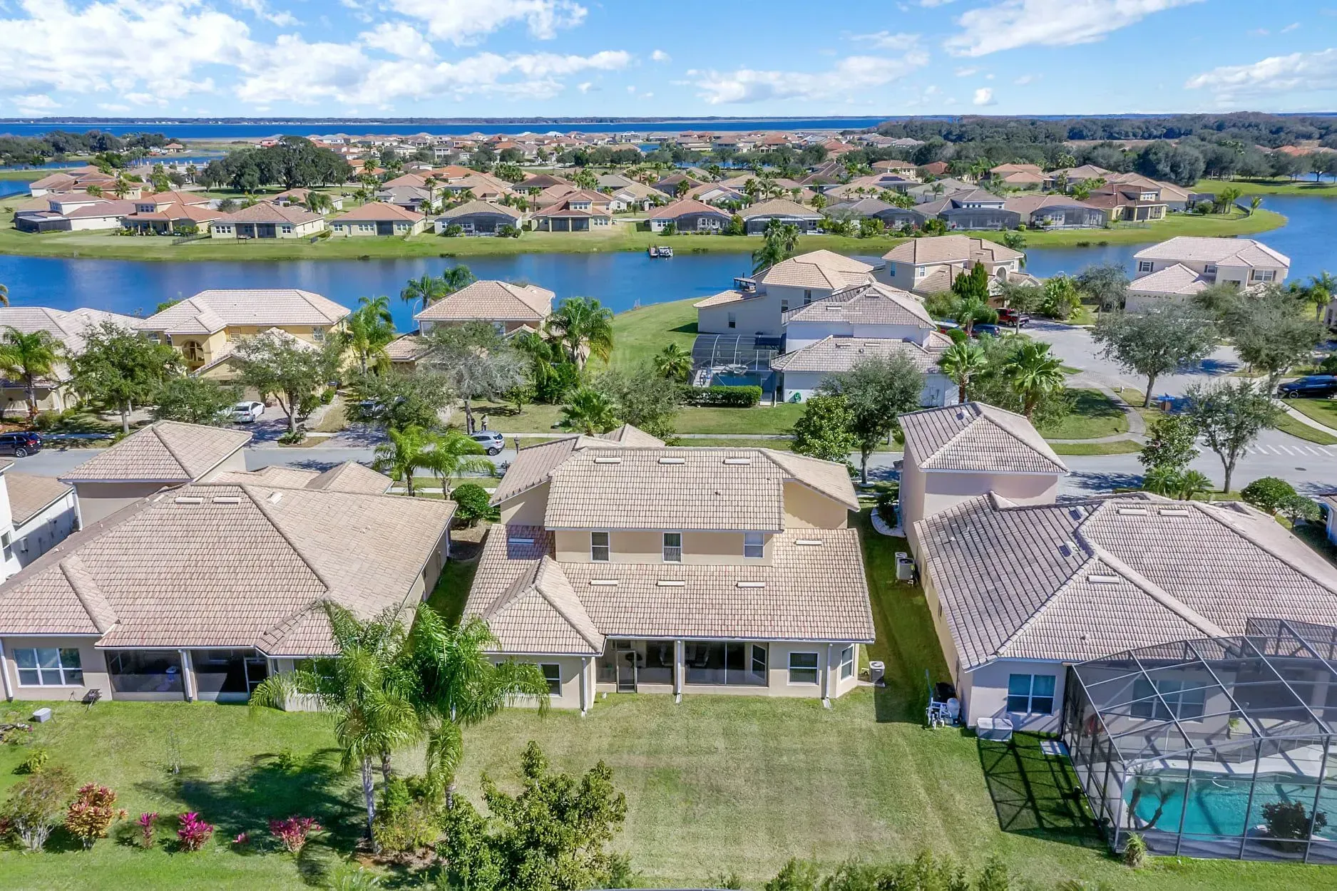 Aerial view of suburban houses with beige tile roofs and a swimming pool near a lake under a sunny blue sky.