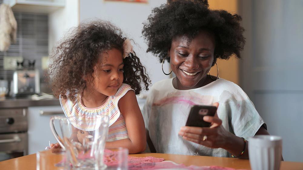 A woman and a little girl are sitting at a table looking at a cell phone.
