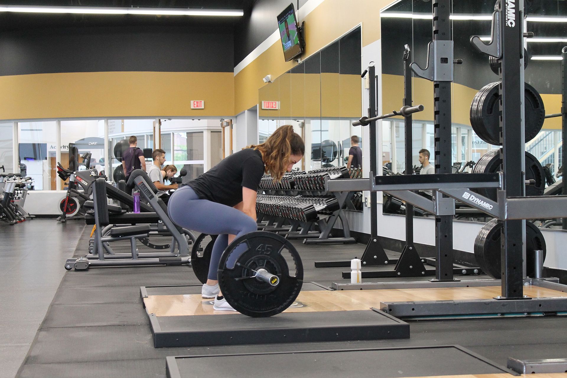 Woman lifting barbell in a gym. She is bent over, lifting, with gray leggings and black shirt.