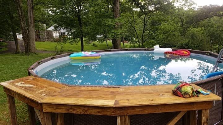 Backyard above-ground pool with toys on a wooden deck, surrounded by trees and grass.