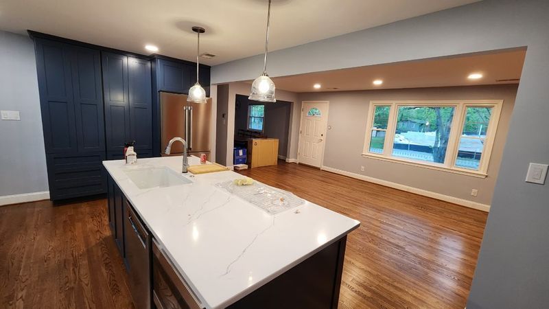 Modern kitchen with white island, dark blue cabinets, hardwood floors, and large windows.