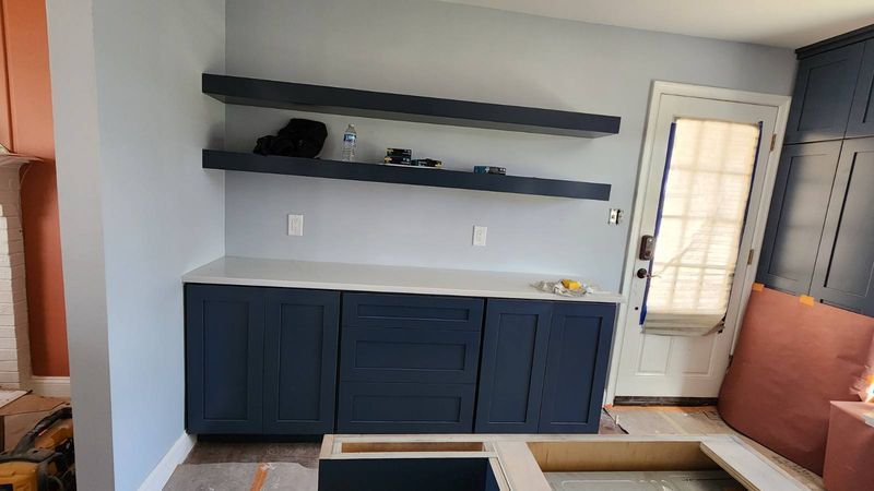 Built-in navy cabinets and floating shelves on a gray wall beside a white door in a kitchen nook.