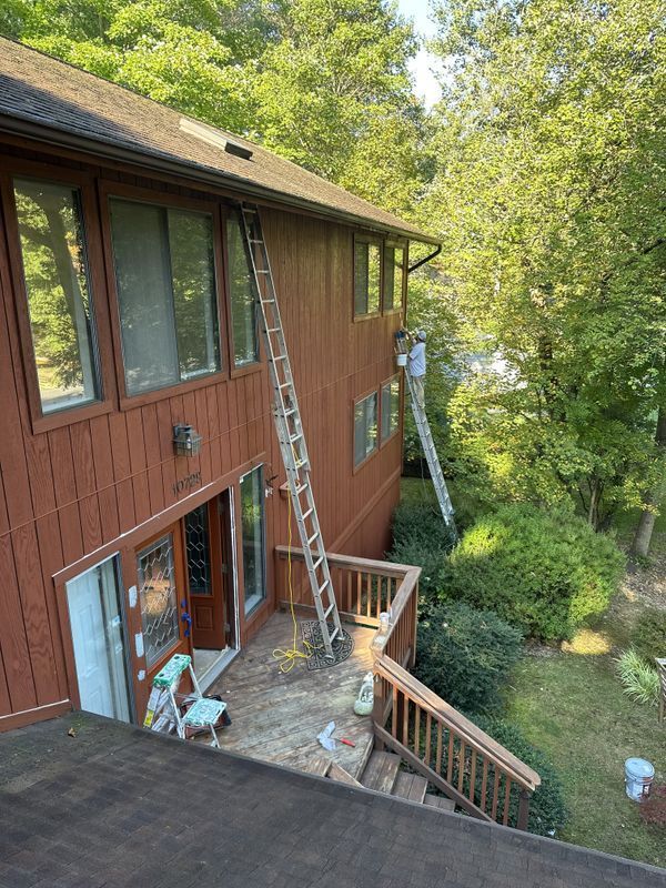 Two-story brown house with ladders on a deck, surrounded by trees and a wooded yard.