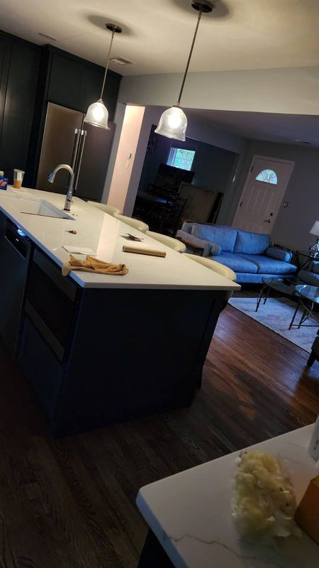 Modern kitchen with island, pendant lights, dark cabinets, and a view into a living room with a couch.