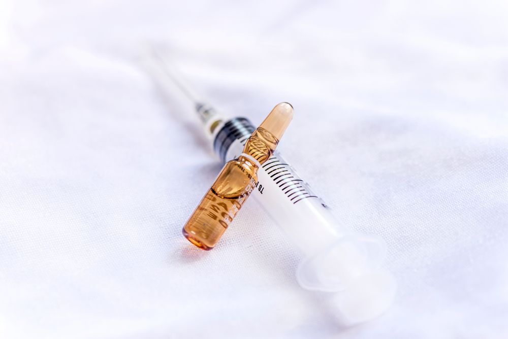 A small, amber-colored medicine ampoule resting diagonally on top of a clear plastic syringe against a white background.
