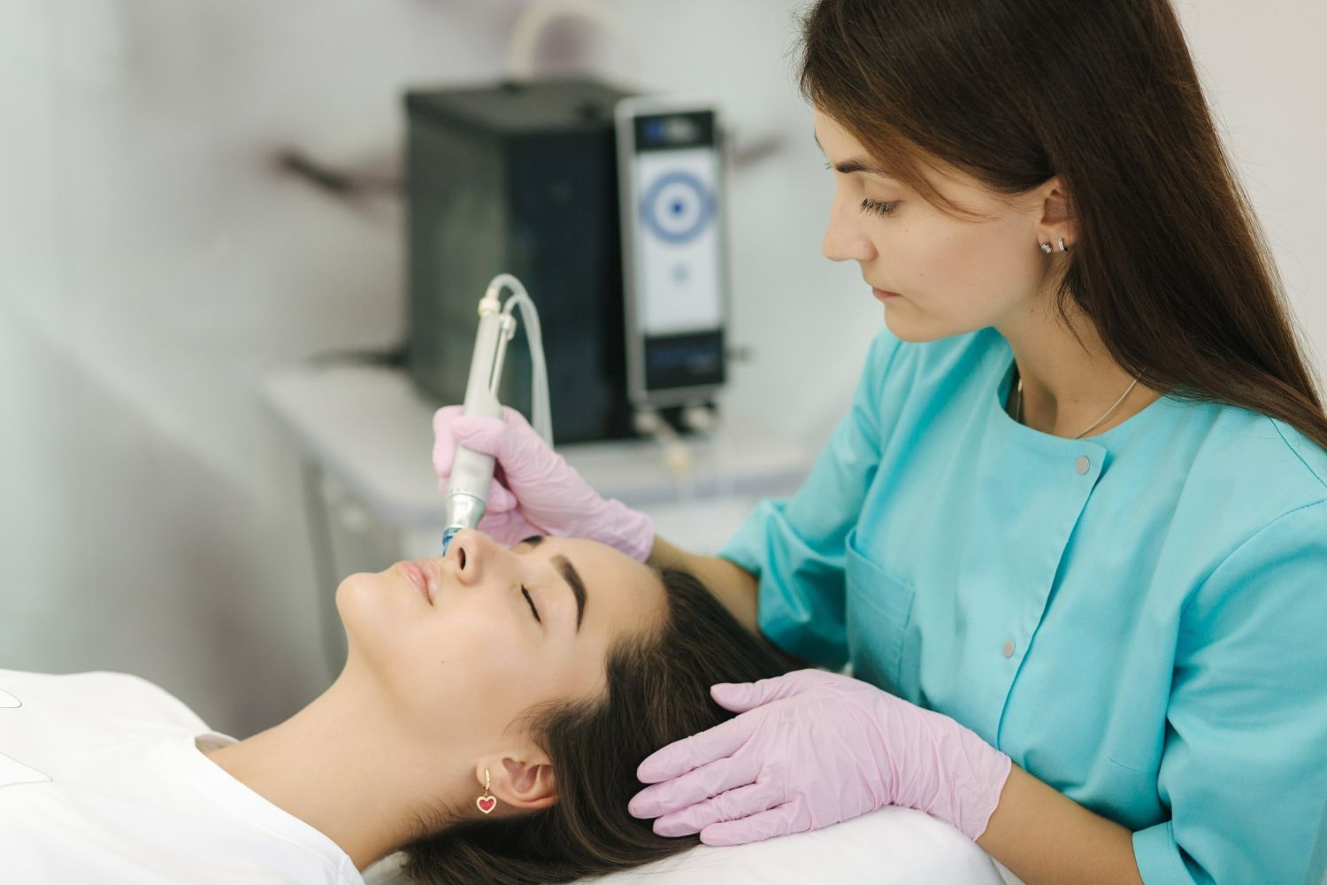 A specialist in a teal scrub top uses a handheld device on a client's face during a skin treatment in a clinical setting.