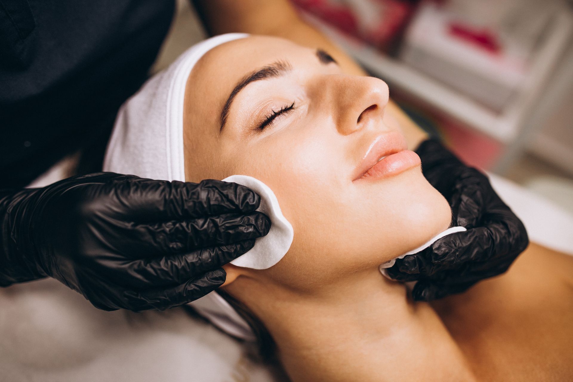 Gloved hands wiping a woman’s face with cotton pads during a facial treatment