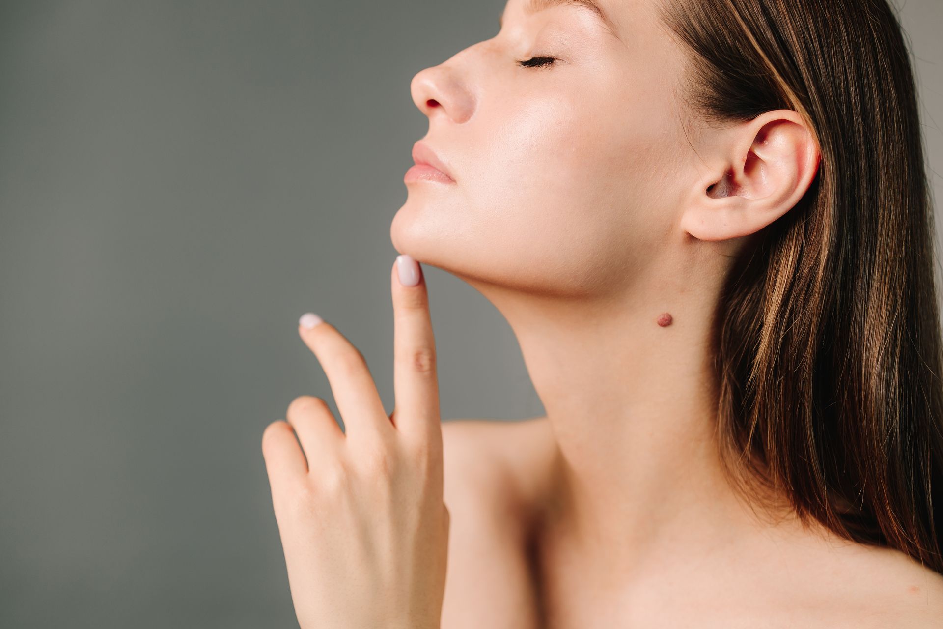 Woman with closed eyes touches her chin; a mole is visible on her neck, gray background.