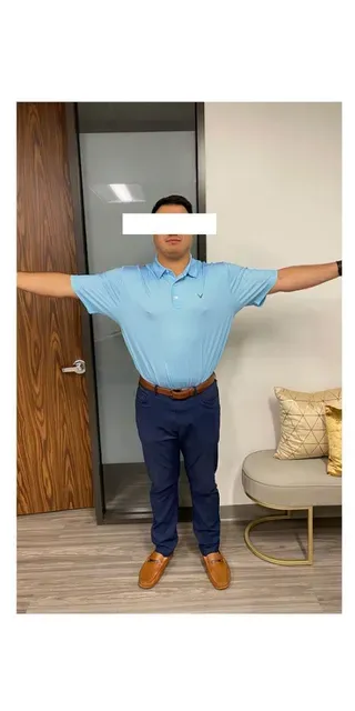 Person with arms outstretched, wearing a blue polo shirt, navy pants, and brown shoes, standing in a room.
