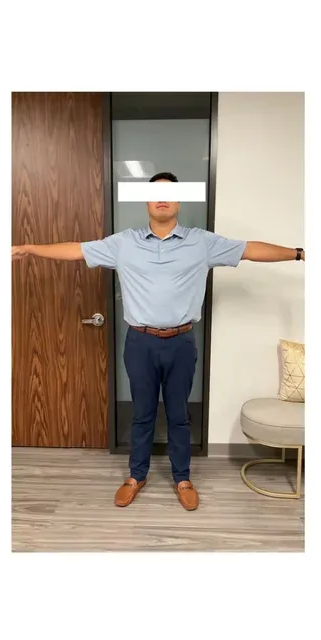 Person stands with arms outstretched in front of a wood door and white wall. They wear a blue polo, pants, and brown shoes.