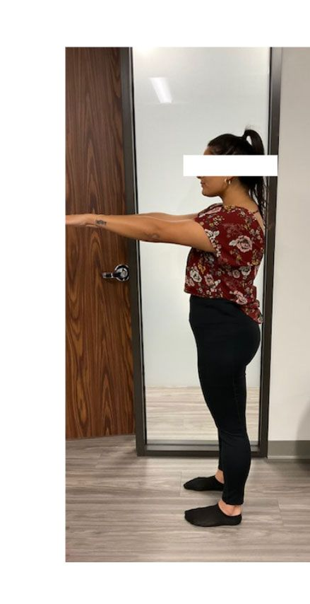 Woman standing sideways with arms outstretched in front of a mirror, wearing black pants and a floral top.