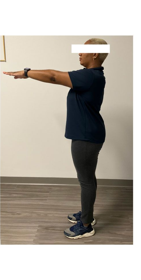 Person in a dark blue shirt and gray pants with arms outstretched, standing in front of a white wall.