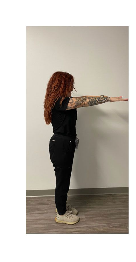 Person in black scrubs, arms outstretched horizontally, standing against a white wall and wooden floor.