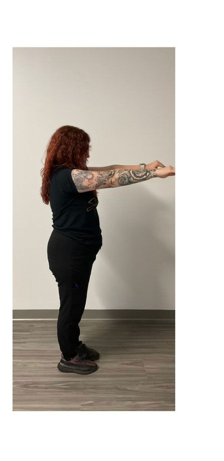 A person with arm tattoos, wearing a black shirt and pants, stands with arms outstretched in front of a white wall.