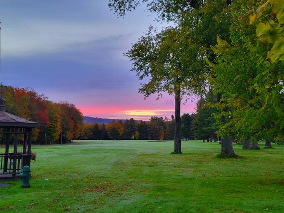 A sunrise glows pink and orange above a green field with trees and a gazebo, creating a peaceful, autumnal park landscape.