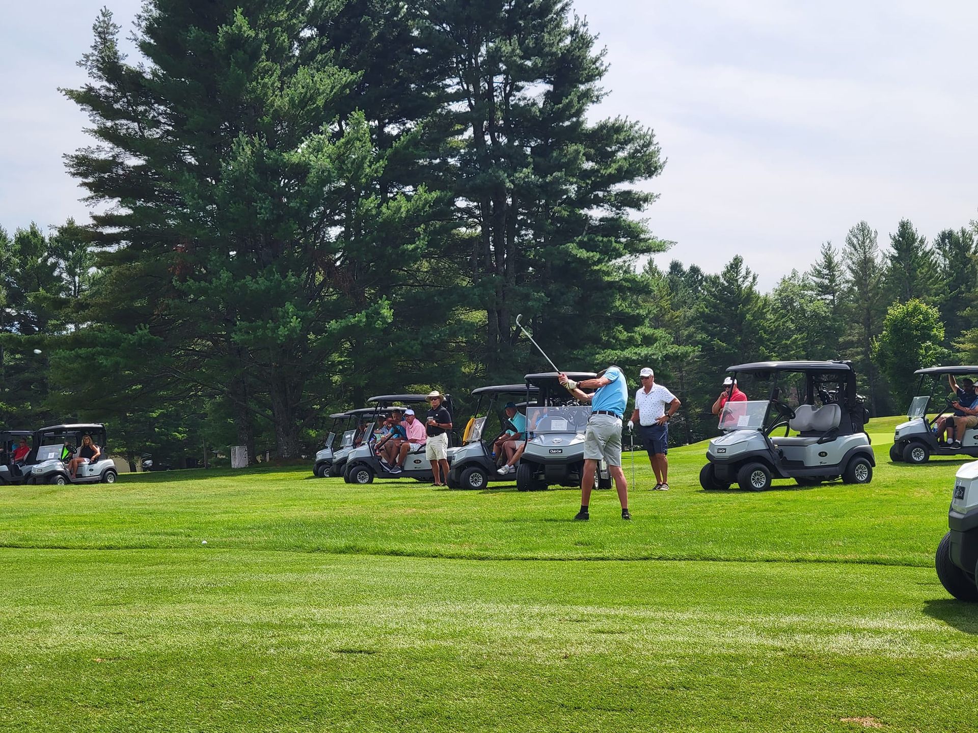A golfer swings a club on a grassy course with several golf carts parked nearby under large, leafy trees.
