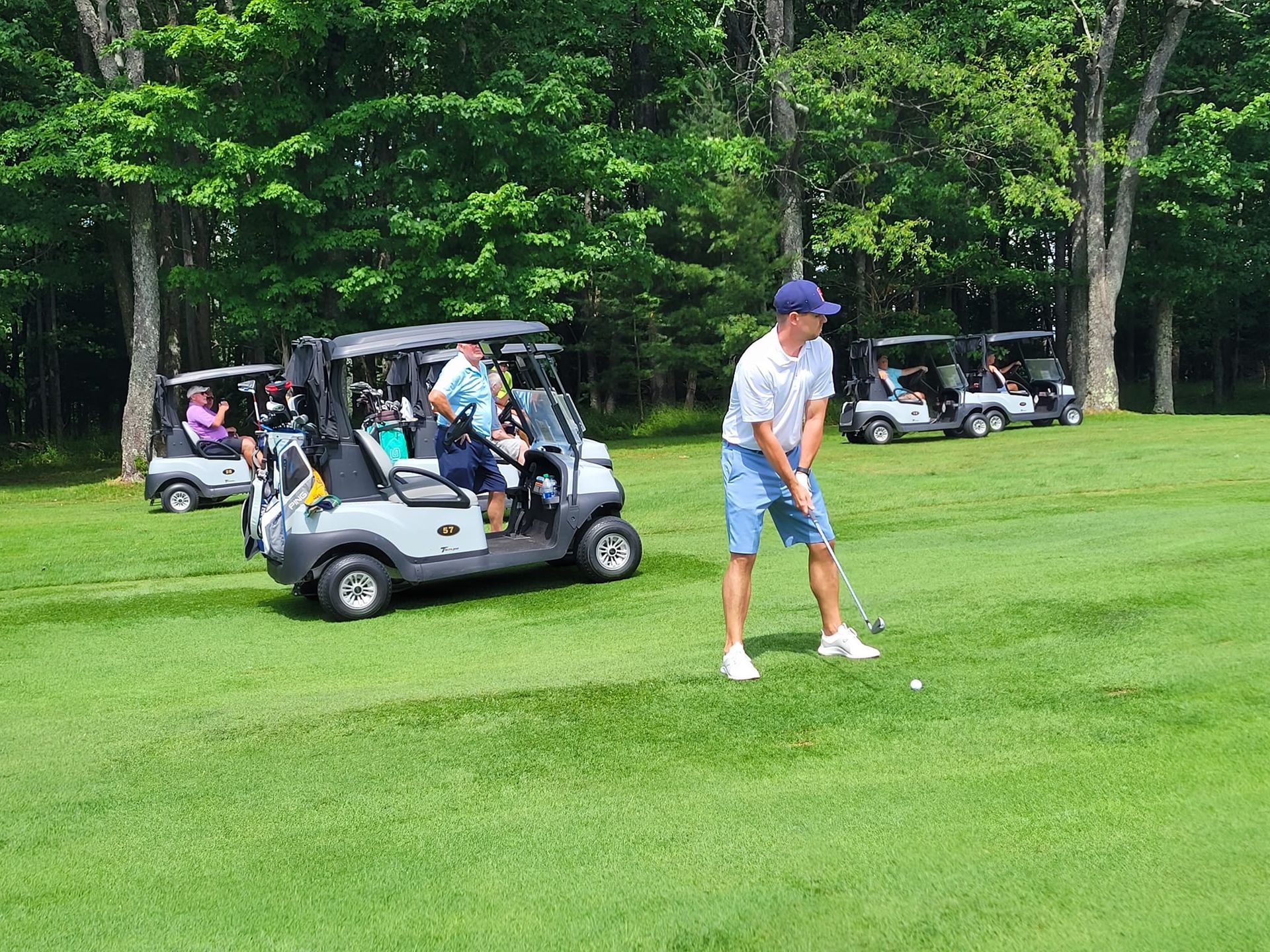 A golfer in a white shirt and blue shorts prepares to swing on a lush green course near several golf carts.