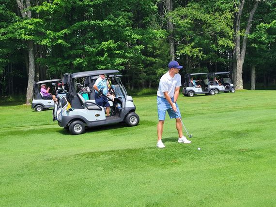 A golfer in a white polo and blue shorts prepares to hit a ball on a green course, with golf carts parked nearby.
