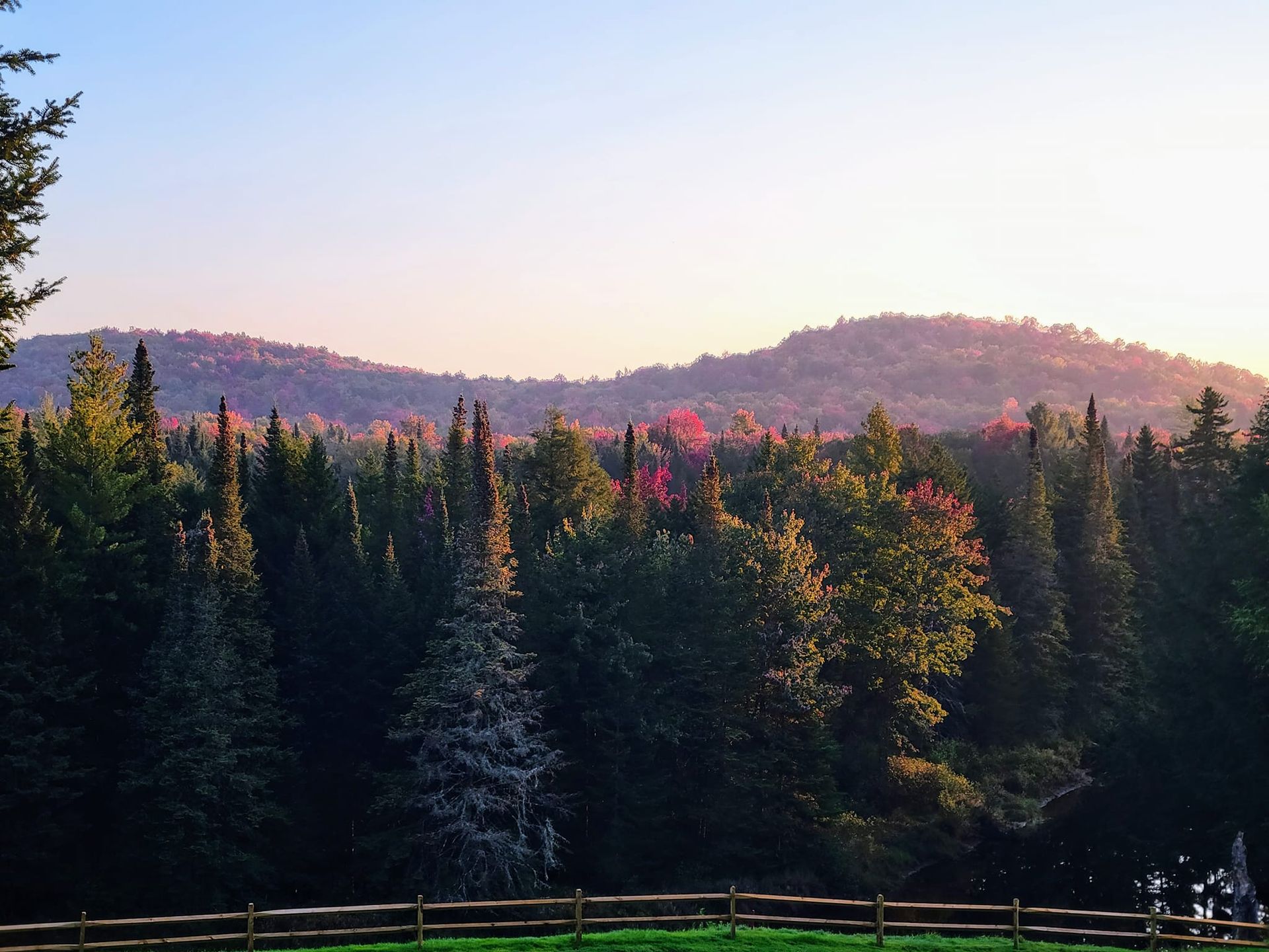 A wooden fence borders a lush forest with tall trees, set against a backdrop of rolling hills under a soft sunset sky.