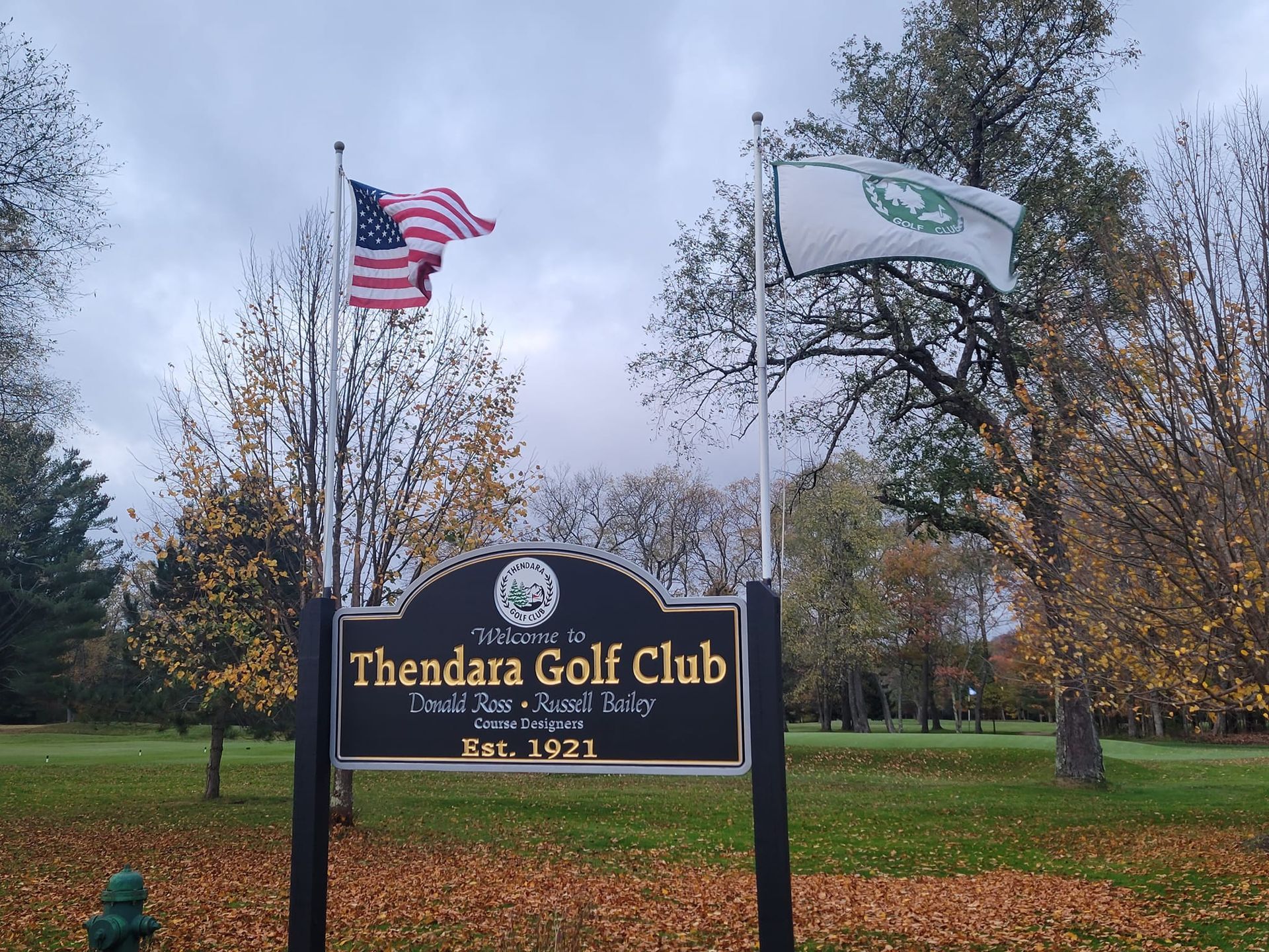 A sign for Thendara Golf Club, established 1921, flanked by an American flag and a white flag on a grassy, autumn-leafed lot.