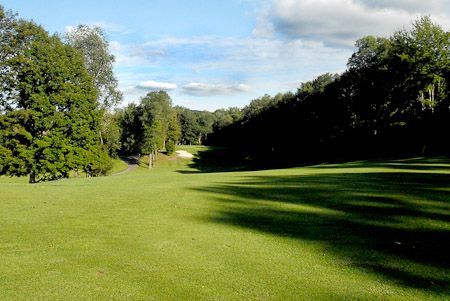 A green golf course fairway slopes downhill toward a distant sand bunker, framed by trees under a bright blue sky.